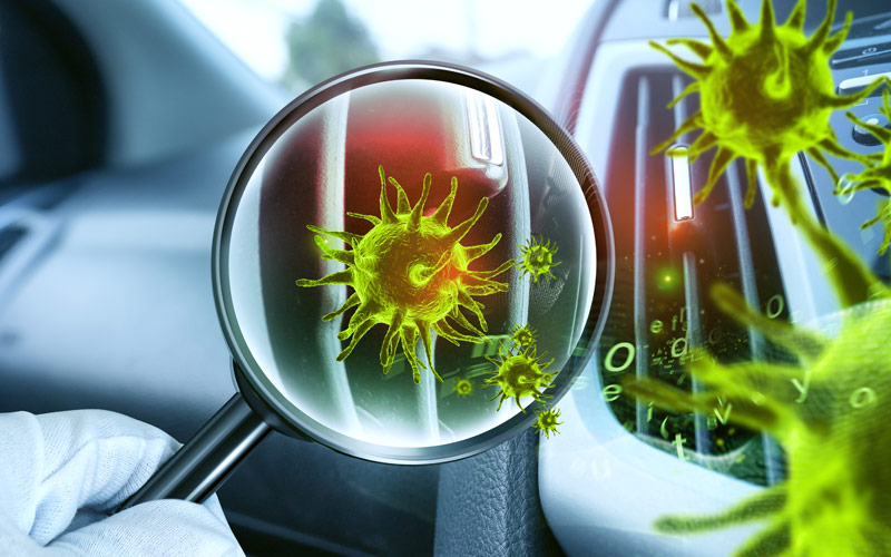 Unter einer Lupe vergrößerte, leuchtend grüne Viren vor dem Lüftungsauslass eines Autos